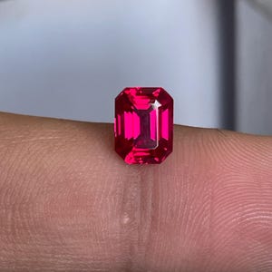May include: A vibrant, deep red gemstone cut in a rectangular emerald shape with faceted surfaces that reflect light. The stone displays a saturated crimson hue and clear internal structure, showcasing precise geometric lines and sharp corners.