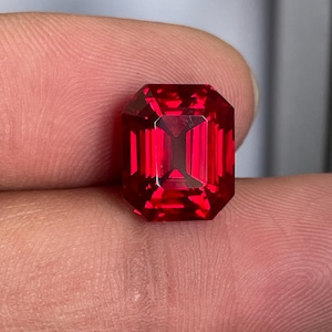 May include: A vibrant, deep red gemstone cut into a rectangular emerald shape with faceted edges. The stone displays high clarity and intense saturation, reflecting light through its geometric facets to create a brilliant, polished appearance.