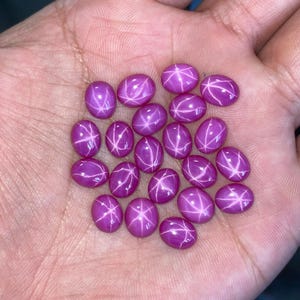 May include: A collection of oval-shaped, magenta-colored gemstones with a star-like effect. The stones are smooth and polished, with a bright, reflective surface. The star effect is created by intersecting lines across the surface of each stone. These gemstones are ideal for jewelry making.