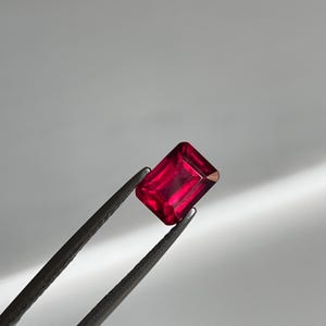 May include: A rectangular-cut ruby gemstone, held by tweezers. The ruby is a deep red colour with a polished surface, reflecting light. The tweezers are dark, contrasting with the gem's brilliance. The background is a soft, neutral grey.