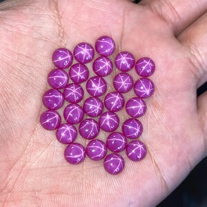 May include: A collection of round, polished, magenta-colored gemstones with a star-like pattern. The gems are displayed in a hand, showcasing their vibrant color and unique optical effect. These gemstones are ideal for jewelry making.