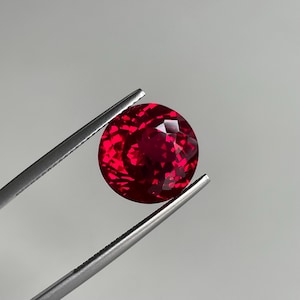 May include: A round, faceted, deep red gemstone held by silver-toned metal tweezers. The stone features a brilliant cut with multiple geometric facets that reflect light, creating a vibrant, saturated crimson appearance against a neutral background.