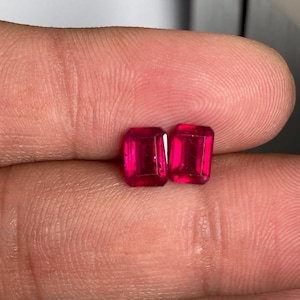 Puede incluir: Dos piedras preciosas de color rojo intenso y vibrante con forma de corte esmeralda. Estas piedras facetadas presentan una silueta rectangular con esquinas afiladas y una superficie pulida que refleja la luz, mostrando un color carmesí rico.