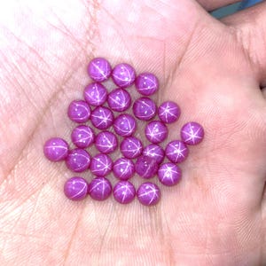 May include: A collection of round, polished, magenta-colored gemstones with a star-like pattern. The gems are held in a hand, showcasing their vibrant color and unique optical effect. These gemstones are ideal for jewelry making.