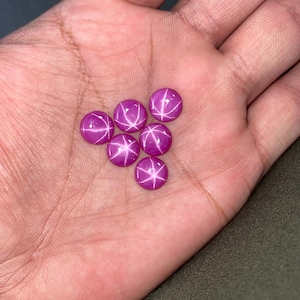 May include: Six small, round, cabochon-cut pink gemstones, each displaying a distinct six-rayed star pattern known as asterism. The stones are smooth, polished, and exhibit a vibrant magenta hue with bright white light reflections forming the star.