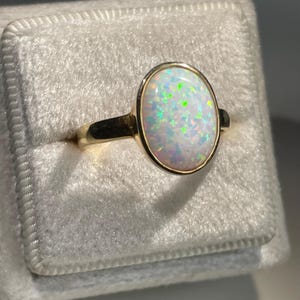 May include: A gold ring featuring an oval-shaped opal gemstone set in a smooth bezel setting. The opal displays a white base color with iridescent flashes of green, blue, and pink throughout the stone. The band is a polished, solid gold metal.