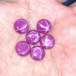 Könnte beinhalten: Sechs runde, polierte Sternrubin-Edelsteine. Jeder Edelstein hat eine tiefe magenta Farbe mit einem sechsstrahligen Sterneffekt. Die Edelsteine werden auf einer hellrosa Oberfläche präsentiert.