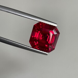 May include: A vibrant, deep red gemstone with a square emerald cut, featuring sharp, clean facets that reflect light. The stone is held securely by a pair of silver-toned metal tweezers, showcasing its intense color and geometric clarity.