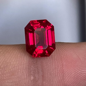 Puede incluir: Una gema de color rojo intenso y vibrante con un corte esmeralda, que presenta múltiples facetas que reflejan la luz para crear un aspecto brillante y centelleante. La piedra tiene un contorno rectangular con esquinas cortadas y una calidad transparente.