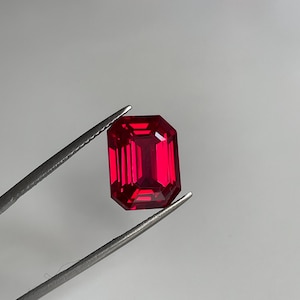 May include: A vibrant, deep red gemstone with an emerald cut, featuring sharp, rectangular facets that reflect light. The gemstone is held securely by a pair of silver-toned metal tweezers, showcasing its clear, saturated color and geometric shape.