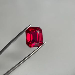 Puede incluir: Una gema de color rojo intenso y vibrante con forma de corte esmeralda, que presenta facetas nítidas y limpias que reflejan la luz. La piedra rectangular está sujeta de forma segura por unas pinzas de metal plateado, mostrando su claridad.