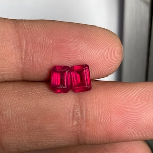 Puede incluir: Dos piedras preciosas de color rojo intenso y vibrante, talladas en forma de esmeralda rectangular. Las piedras presentan superficies facetadas que reflejan la luz, mostrando un tono carmesí saturado y bordes claros y definidos.