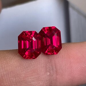 May include: Two vibrant red gemstones with an emerald cut, featuring a rectangular shape and faceted surfaces that reflect light. These loose stones display a deep, saturated crimson hue and are positioned side by side to showcase their clarity.