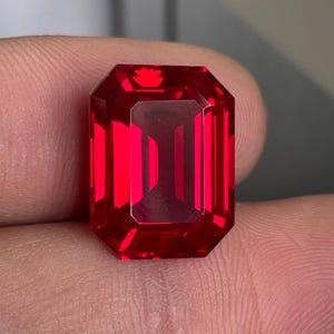Op de afbeelding: Een levendige, dieprode edelsteen geslepen in een rechthoekige smaragdvorm met scherpe, heldere facetten. De steen vertoont een hoge mate van helderheid en intense verzadiging, waarbij het licht door de geometrische structuur wordt weerkaatst.