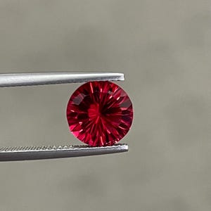 Può includere: Una gemma rotonda di colore rosso vibrante con un motivo sfaccettato a forma di stella che irradia dal centro. La gemma è tenuta saldamente tra le due punte metalliche di una pinzetta color argento, mettendo in risalto il suo colore cremisi.