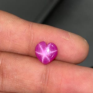 May include: A heart-shaped star ruby gemstone featuring a vibrant magenta-pink hue and a distinct six-rayed star pattern visible on its polished surface. The gemstone displays a smooth, rounded contour and a reflective, translucent quality.