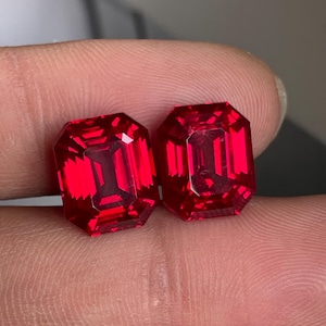 May include: Two vibrant red gemstones with an emerald cut, featuring a rectangular shape and faceted surfaces that reflect light. The stones are clear and display a deep, saturated crimson hue throughout their structure, showcasing precise geometric edges.