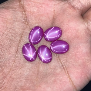 May include: Five oval-shaped, cabochon-cut gemstones with a vibrant purple hue. Each stone displays a distinct, bright white star-shaped optical phenomenon known as asterism, centered on the smooth, polished, and rounded surface of the gems.