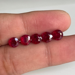 May include: Five round, faceted, deep red rubies are displayed. The gemstones are arranged in a row, showcasing their brilliant cut and rich color. The rubies are set against a neutral background, highlighting their vibrant hue and clarity.