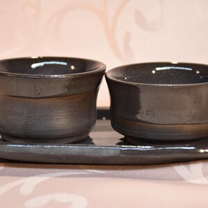 May include: A pair of dark grey ceramic cups on a matching oval tray. The cups have a slightly flared rim and a dark, glossy finish. The tray is also dark grey and has a smooth, reflective surface. The set appears to be handmade.