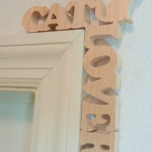 May include: A wooden scroll saw pattern reading "CAT LOVER" with a cat silhouette. The text is in capital letters and is positioned at a right angle. The words are in a light wood color and are mounted on a white wall.