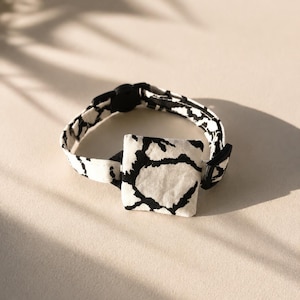 May include: A black and white patterned pet collar with a square accent. The collar features a black buckle closure and a repeating abstract design on a white background. The collar is designed for a small pet.
