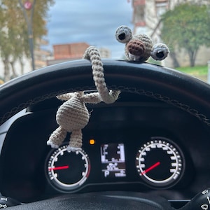 Pode incluir: Um pendente de carro de macaco de crochê, bege e castanho, pendurado no volante de um carro. O macaco tem olhos grandes e redondos e um rosto sorridente. O painel e os indicadores do carro são visíveis no fundo.