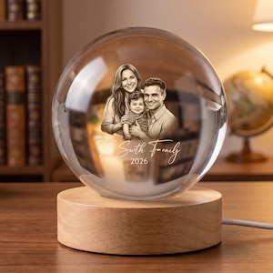 May include: A clear glass sphere resting on a cylindrical light-colored wooden base. The sphere features an etched monochrome portrait of a family of three with the text "Smith Family 2026" engraved below the figures. A white power cord extends from the base.