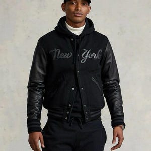 May include: Black varsity jacket with leather sleeves and the words "New York" embroidered across the chest. The jacket has a button-up front and ribbed cuffs. A black baseball cap with a logo is also visible.