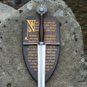 May include: A decorative sword with a silver blade and a dark brown handle, resting on a large stone. The sword is mounted on a wooden plaque with gold lettering. The plaque features an ornate initial and text.