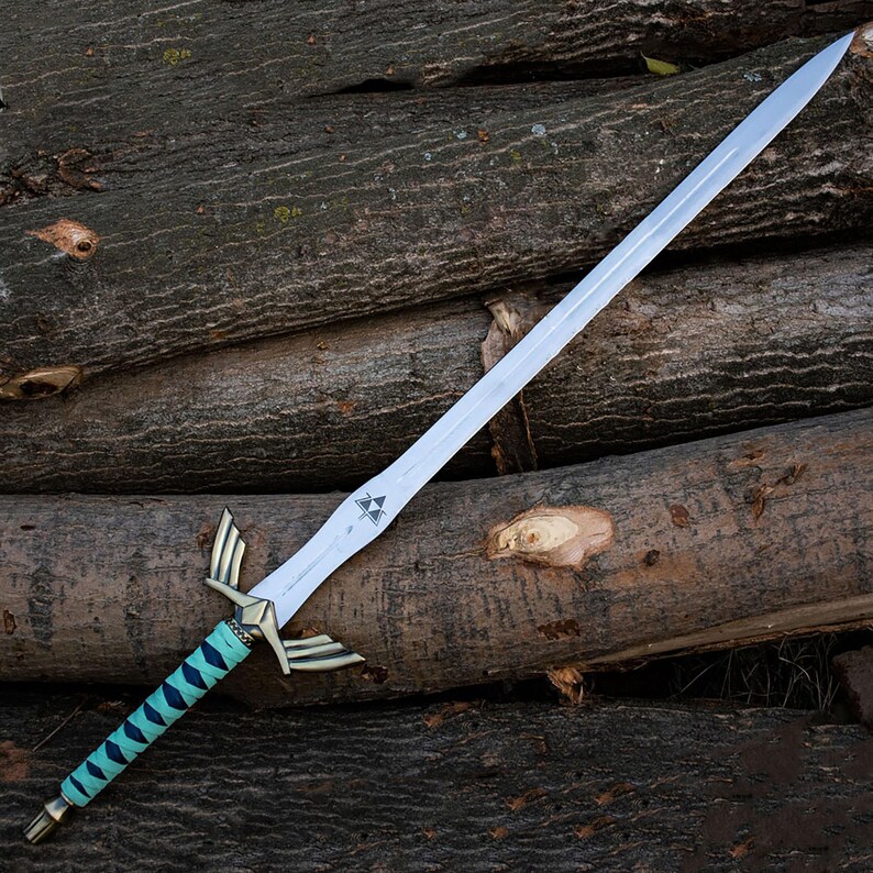 Peut inclure: Une longue &eacute;p&eacute;e en m&eacute;tal dot&eacute;e d'une lame argent&eacute;e avec un petit embl&egrave;me triangulaire pr&egrave;s de la garde. La poign&eacute;e pr&eacute;sente une garde en bronze avec des extensions en forme d'ailes et un manche envelopp&eacute; de turquoise vif et bleu fonc&eacute;.