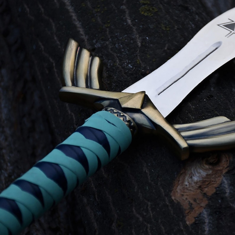 Peut inclure: Une &eacute;p&eacute;e avec une lame en argent m&eacute;tallique pr&eacute;sentant un symbole triangulaire pr&egrave;s de la garde. La poign&eacute;e est en laiton antique avec des gardes en forme d'ailes et un manche envelopp&eacute; de bandes altern&eacute;es de cuir bleu fonc&eacute; et bleu sarcelle.