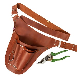 Op de afbeelding: Een bruine leren tuingereedschapsriem met meerdere vakken en een schouderriem. Inclusief een snoeischaar met groene handgreep. De riem heeft een metalen ring en klinknagels.