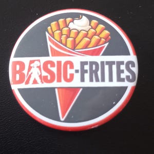 Peut inclure: Un badge rond épinglette illustré d'un cornet rouge rempli de frites dorées surmontées d'une noisette de mayonnaise blanche. Le texte BASIC-FRITES est imprimé en lettres rouges et noires audacieuses au centre du motif circulaire.