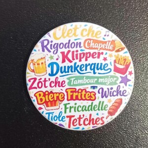 Peut inclure: Un badge rond avec du texte coloré incluant Clet'che, Rigodon, Chapelle, Klipper, Dunkerque, Zot'che, Tambour major, Biere, Frites, Wiche, Fricadelle, Tiole et Tet'ches, accompagné d'illustrations d'une chope de bière, de frites et d'une saucisse.