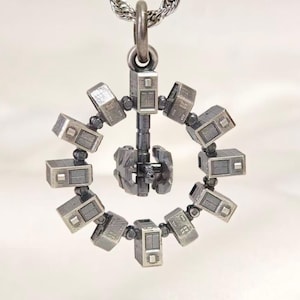 May include: A metallic silver-toned pendant shaped like a rotating space station. The design features a central hub connected by a vertical strut to an outer ring composed of ten rectangular modules, all suspended from a silver chain link.