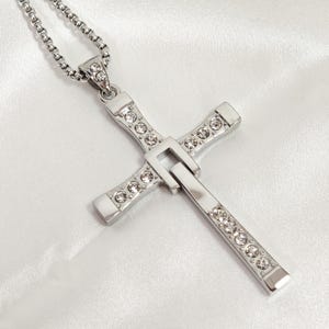 Puede incluir: Collar con colgante de cruz plateada y cadena fina. La cruz está adornada con cristales transparentes. El collar se muestra sobre un fondo blanco texturizado.