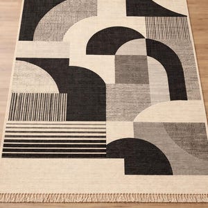 May include: Geometric patterned area rug in black, gray, and cream. The rug features a repeating design of arches, rectangles, and stripes. The rug has a fringed edge and is made of a woven material. A modern, stylish home decor item.