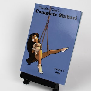 May include: A book titled "Douglas Kent's Complete Shibari: Volume 2 Sky" with an illustration of a woman in a blue sky background.