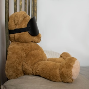Pu&ograve; includere: Un orsacchiotto di peluche marrone con una maschera per dormire nera &egrave; sdraiato su un letto beige. L'orsacchiotto &egrave; rivolto a destra e ha gli occhi chiusi.