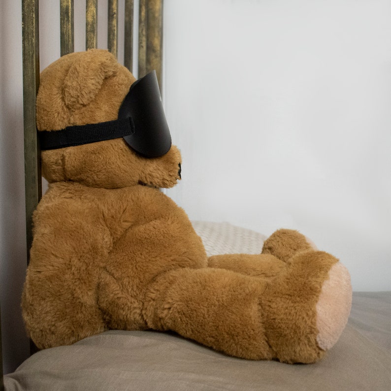 Pu&ograve; includere: Un orsacchiotto di peluche marrone con una maschera per dormire nera &egrave; sdraiato su un letto beige. L'orsacchiotto &egrave; rivolto a destra e ha gli occhi chiusi.