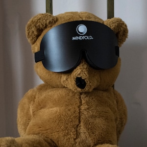 Pu&ograve; includere: Un orsacchiotto di peluche marrone che indossa una maschera per dormire nera con la scritta "MINDFOLD." stampata su di essa.