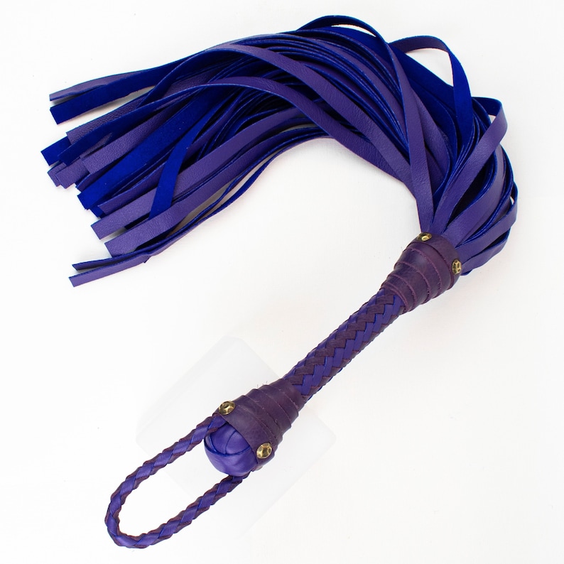 Puede incluir: Un l&aacute;tigo de cuero morado con un mango trenzado y una bola redonda en el extremo. El l&aacute;tigo tiene una franja de tiras de cuero.