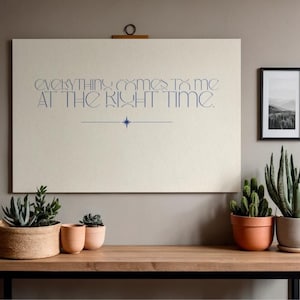 Puede incluir: Un póster grande, blanquecino con el texto "EVERYTHING COMES TO ME AT THE RIGHT TIME" en letras azules. El póster está colgado sobre una mesa de madera con varias plantas en macetas, incluyendo suculentas y cactus. A la derecha hay una foto de paisaje en blanco y negro enmarcada.