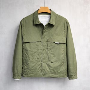 Könnte beinhalten: Olivgrüne Jacke mit Knopfleiste, zwei Brusttaschen und zwei unteren Taschen. Die Jacke hängt an einem Holzbügel. Darunter ist ein weißes T-Shirt zu sehen.