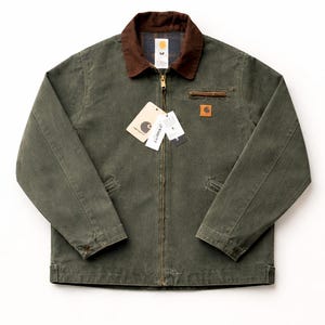 Puede incluir: Chaqueta Carhartt verde con cuello de pana marrón y cremallera. La chaqueta tiene dos bolsillos delanteros y un pequeño parche cuadrado con el logotipo. Varias etiquetas cuelgan de la cremallera.