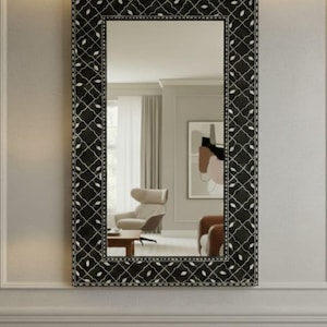 May include: A rectangular wall mirror with a black frame featuring a white geometric pattern. The mirror reflects a room with furniture and artwork. The frame's design adds a decorative element to the mirror.