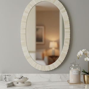 Peut inclure: Miroir ovale avec un cadre festonné blanc cassé. Le miroir est accroché sur un mur gris au-dessus d'un comptoir en marbre. Un pot avec une étiquette et un pot doré avec une plante sont sur le comptoir.