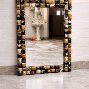 May include: A rectangular mirror with a decorative frame. The frame is made up of small, square tiles in shades of brown, black, and cream. The mirror is propped against a light-coloured wall and floor.