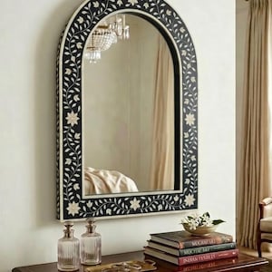 May include: An arched wall mirror with a black frame, adorned with white floral and vine details. The mirror is hanging above a dark wooden table, with books, decorative bottles, and a tray with jewelry. The mirror reflects a chandelier and a bed.
